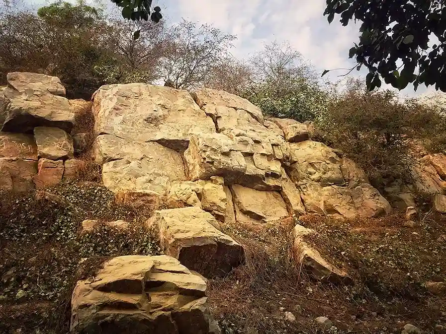 A natural landscape photograph of the sacred Govardhan Hill, showcasing its rugged, light-brown rocky terrain. Large weathered boulders are interspersed with sparse green shrubs and dry trees under a bright, clear sky.