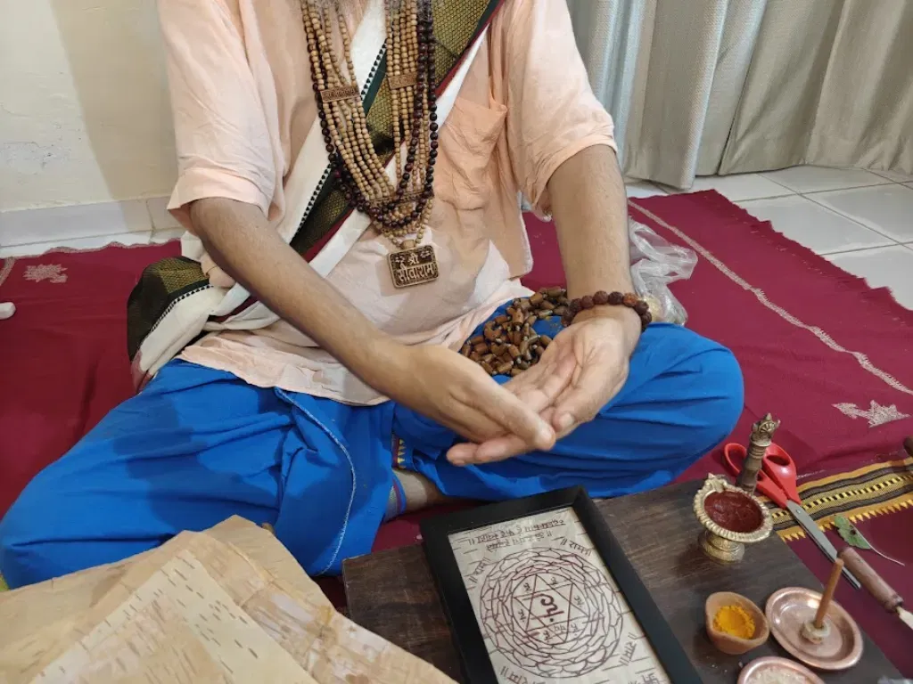A high-angle, medium shot of Shri Damodar Dasji Maharaj seated in a blue dhoti, performing Prana-Pratishtha. His hands are held in a specific energizing Mudra over a hand-drawn Yantra on Himalayan Bhojpatra. The Yantra is framed and placed on a wooden surface alongside traditional Upakaranas, including Akshat, Haldi, and a natural ink diya. Shri Damodar Dasji Maharaj wears multiple sacred malas and a "Sita-Ram" pendant, embodying the Ramanandi Sampradaya lineage.