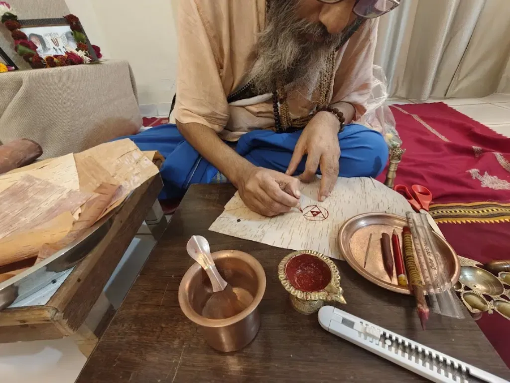 A high-angle, close-up shot of Shri Damodar Dasji Maharaj, a bearded elder in traditional saffron and blue spiritual attire, meticulously hand-drawing a geometric Yantra. He is seated on the floor, using a traditional stylus to mark a piece of authentic Himalayan Bhojpatra (birch bark). On the wooden table before him are sacred tools including a small copper vessel of water, a diya containing red ritual paste, a ruler, and several natural styluses. In the background, a framed sacred image adorned with a flower garland sits on an altar.